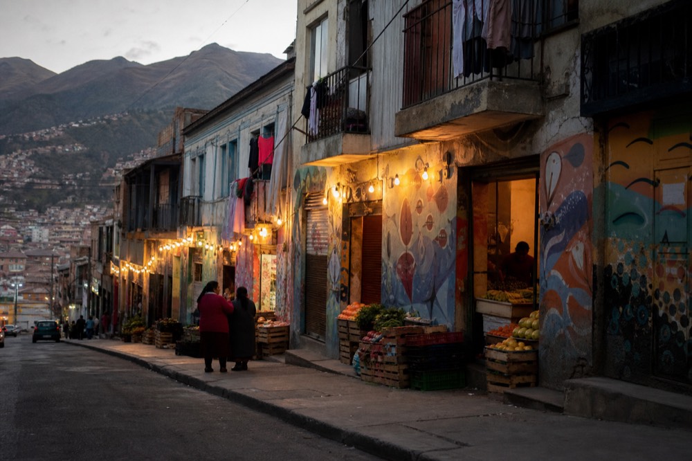 Row of small open shopfronts along a sloping street with fruit crates outside and residents chatting, laundry on balconies above, ordinary weekday light