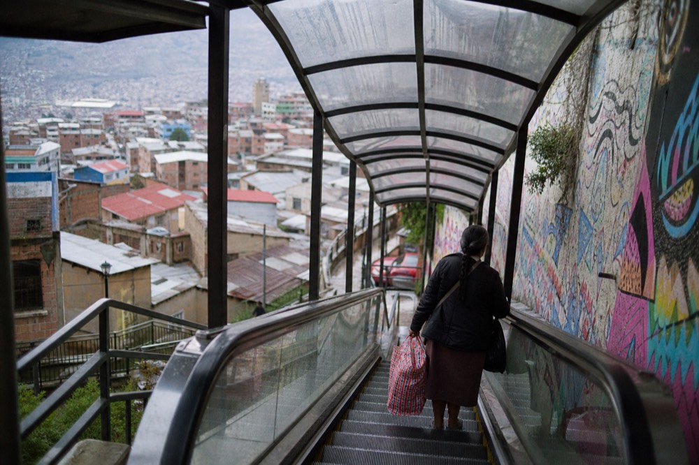 Covered outdoor escalator flight climbing between houses with a woman carrying shopping bags riding up, hillside rooftops falling away behind
