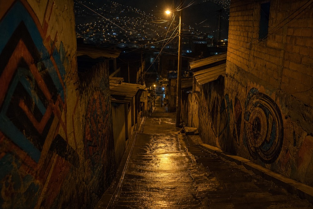 A narrow hillside alley of steps at night lit by a single bare bulb, wet concrete reflecting the light, no people visible