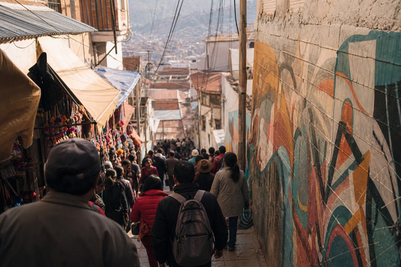 Dense crowd filling a narrow shop-lined hillside walkway seen from above, awnings and hanging merchandise overhead, late morning