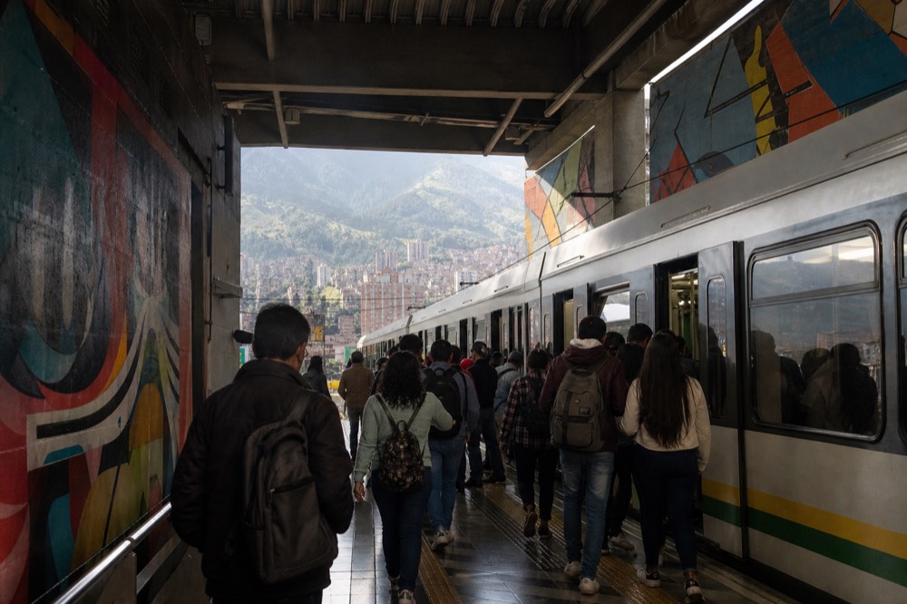 A busy elevated metro platform in a valley city at midday, commuters seen from behind boarding a train, green mountainsides visible through the station opening