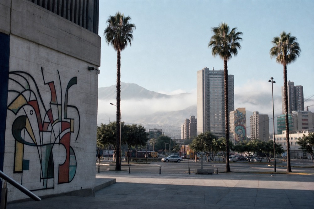 Wide open plaza with tall palms, modern concrete towers and distant figures crossing in bright midday light