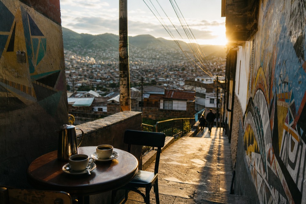 Small terrace cafe table with two cups of black coffee and a metal pot, green hills and low rooftops beyond the railing