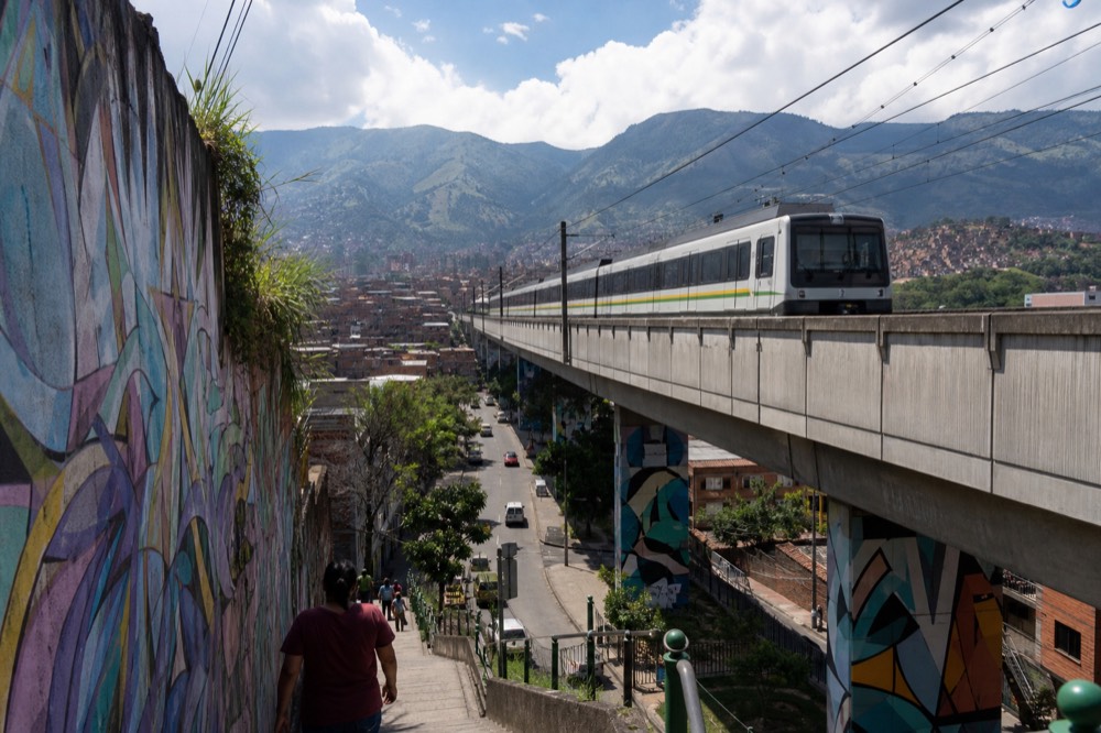 Metro train arriving on an elevated concrete viaduct above city streets, rooftops and green hills stretching behind under bright cloud
