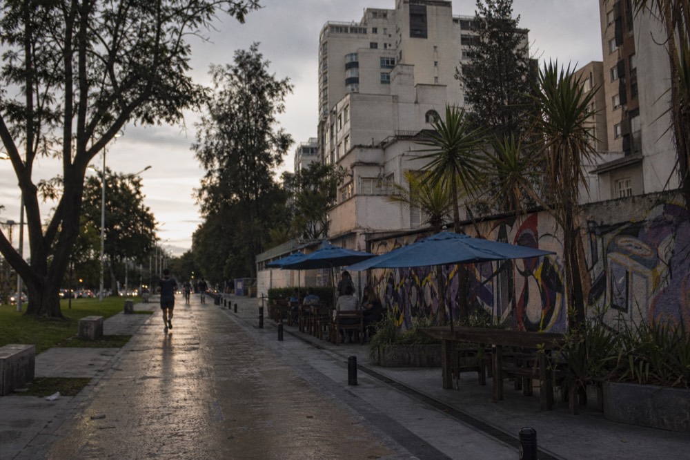 Wide tree-lined pedestrian avenue with joggers and outdoor tables beside low-rise apartment buildings under an early evening sky