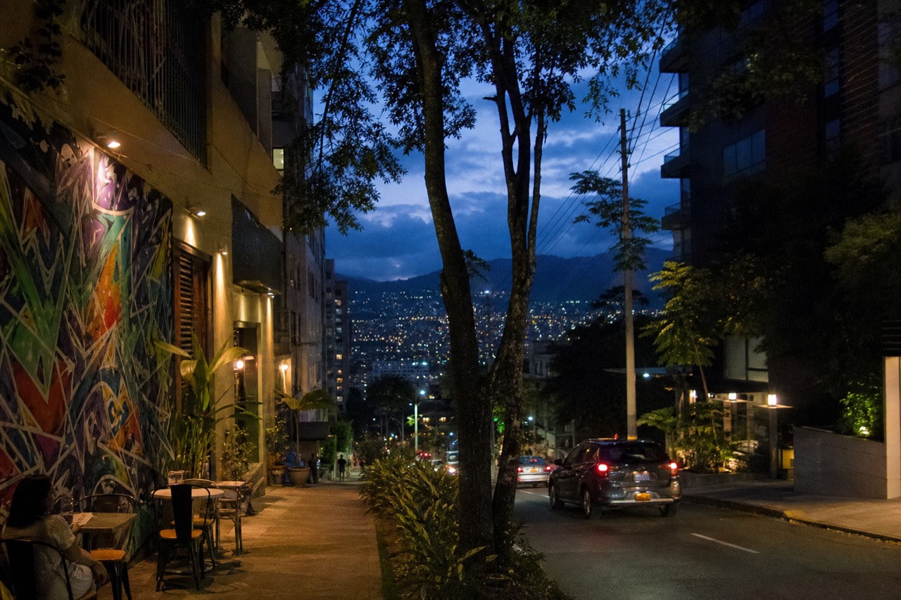 Leafy upscale Medellin street of modern apartment towers and sidewalk cafes at dusk, warm window light and green street trees