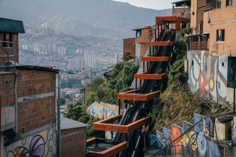 Long flight of outdoor escalators under bright orange canopies climbing a hillside between brick homes, a few people riding up in morning light