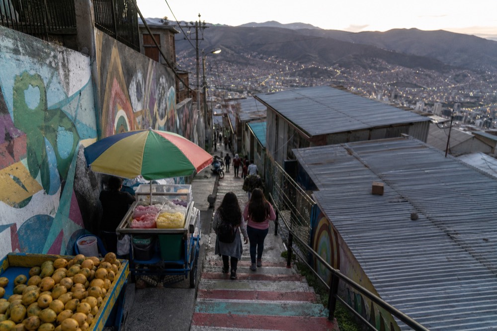 Visitors climbing a bright painted stairway past a mango cart and a shaved-ice stand, seen from above in strong midday light