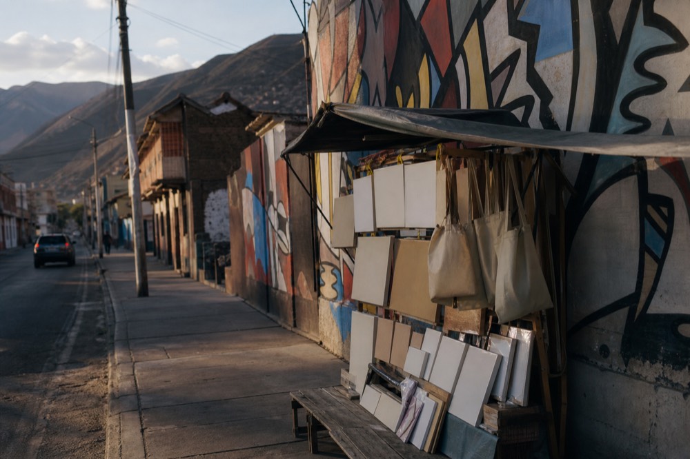 Souvenir stall of small unlabelled canvases and plain tote bags hanging under a tarpaulin beside a painted concrete wall
