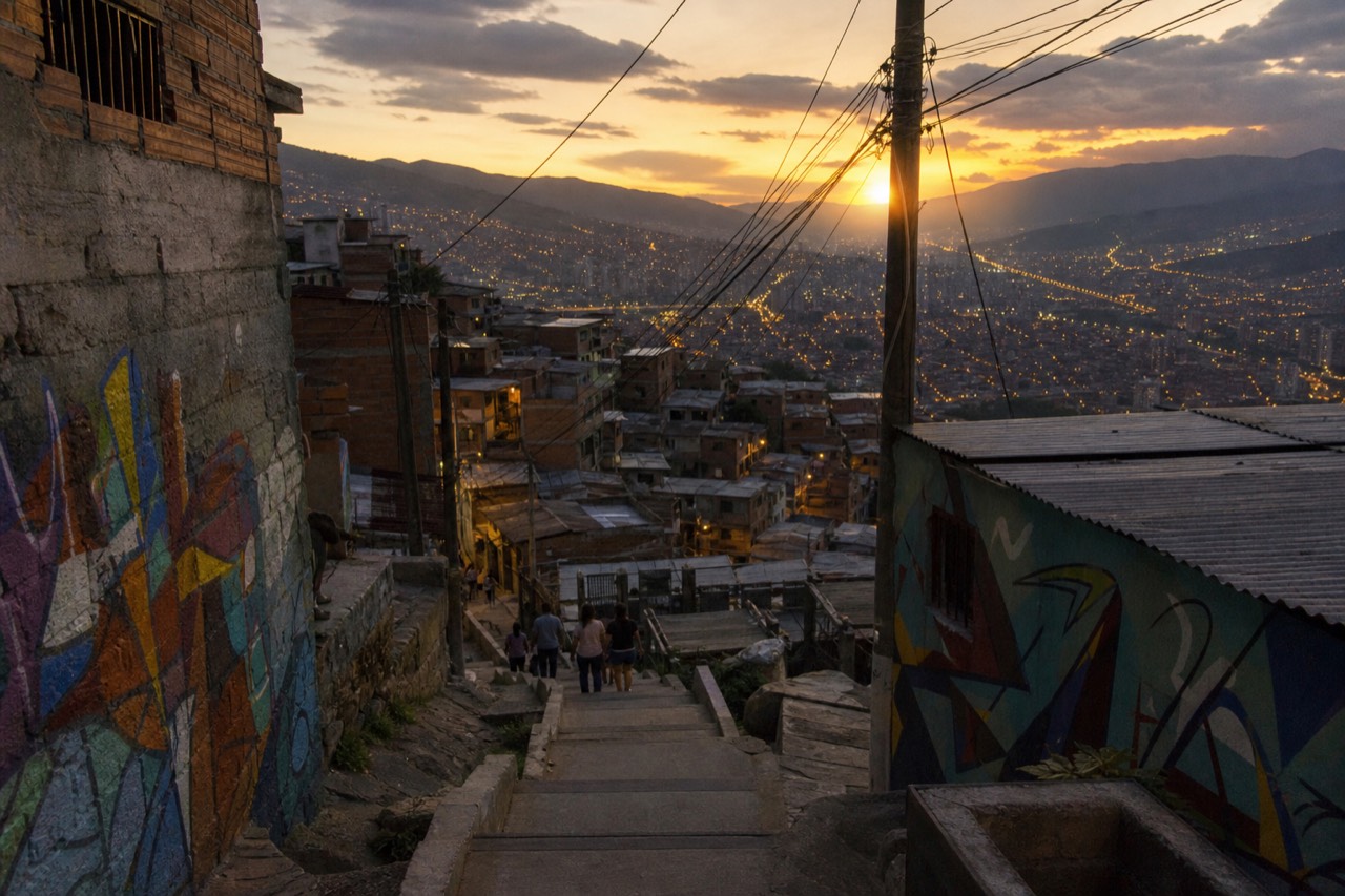 Panorama of a dense hillside neighborhood in Medellin at sunset, ridges of brick houses stepping down toward valley lights