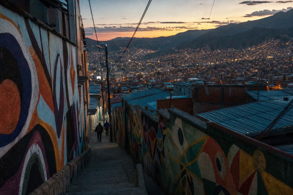 Narrow concrete stairway alley climbing between houses, walls covered in colourful abstract murals, two distant figures walking up