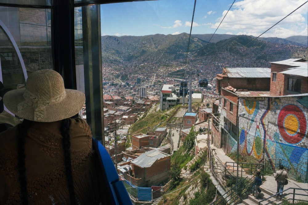 View from inside a cable car cabin over dense hillside rooftops and green ridges, cables and a distant station visible ahead