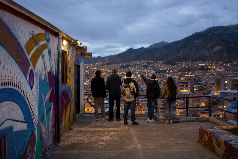 Small group of visitors seen from behind on a concrete terrace overlooking painted rooftops, one person gesturing towards the valley
