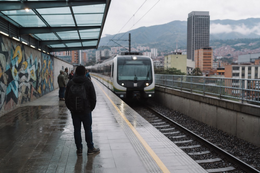 Early morning metro platform in Medellin with a train arriving, a few commuters waiting, tiled floor and glass canopy overhead