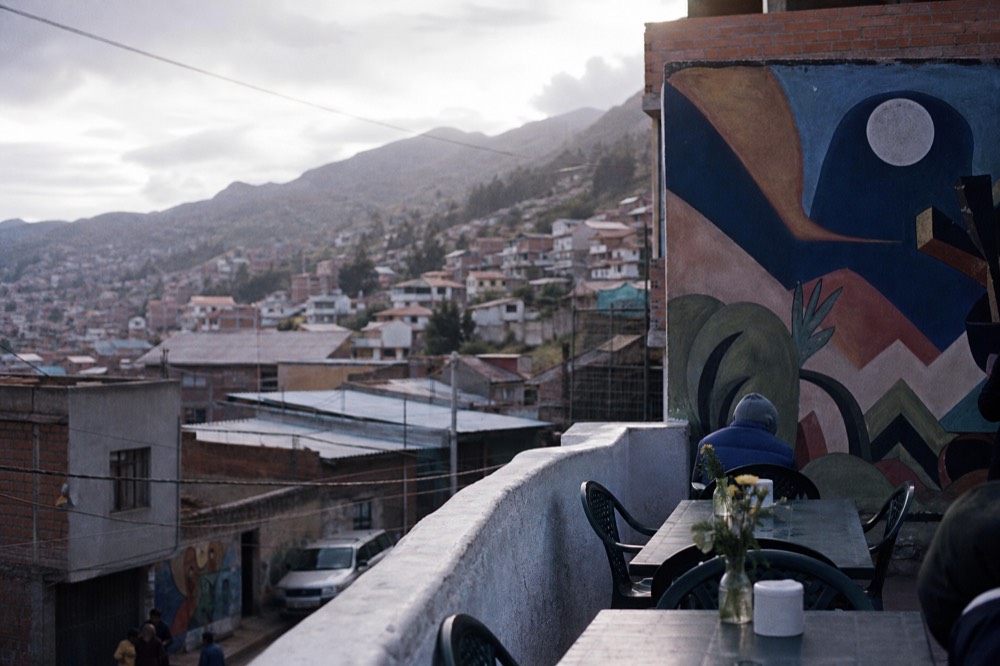 Rooftop terrace cafe with plastic chairs and a low wall, looking out over stacked hillside houses under a bright overcast sky