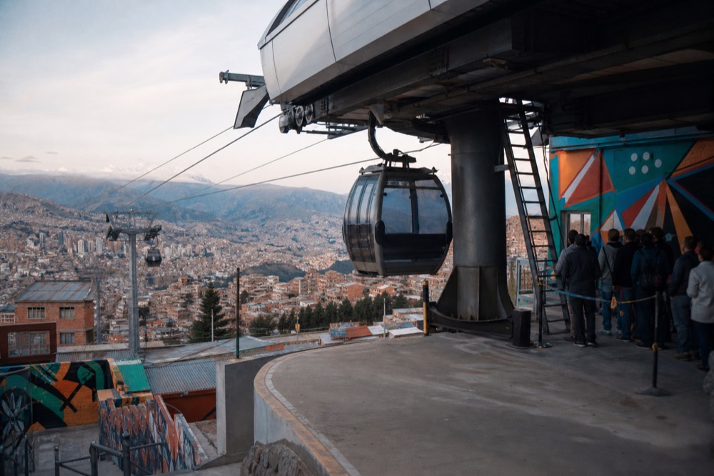 A glass cable-car cabin arriving at an open hillside station in mid-morning light, a short queue of distant passengers waiting on the platform
