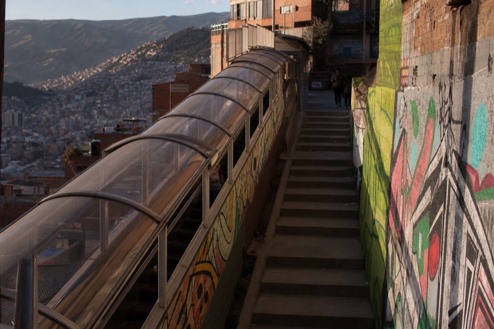 A long covered outdoor escalator section climbing a steep hillside beside a parallel flight of concrete stairs, brick houses tight on both sides