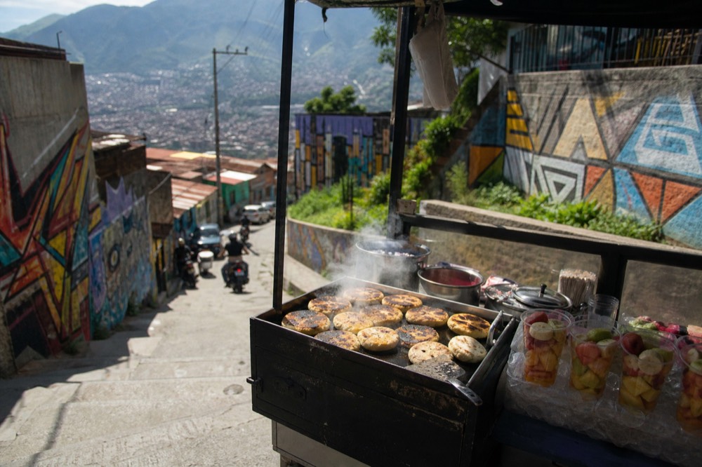 A street food stall on a hillside landing at mid-morning, arepas on a hot griddle and fresh fruit cups lined up, steam rising, no faces visible