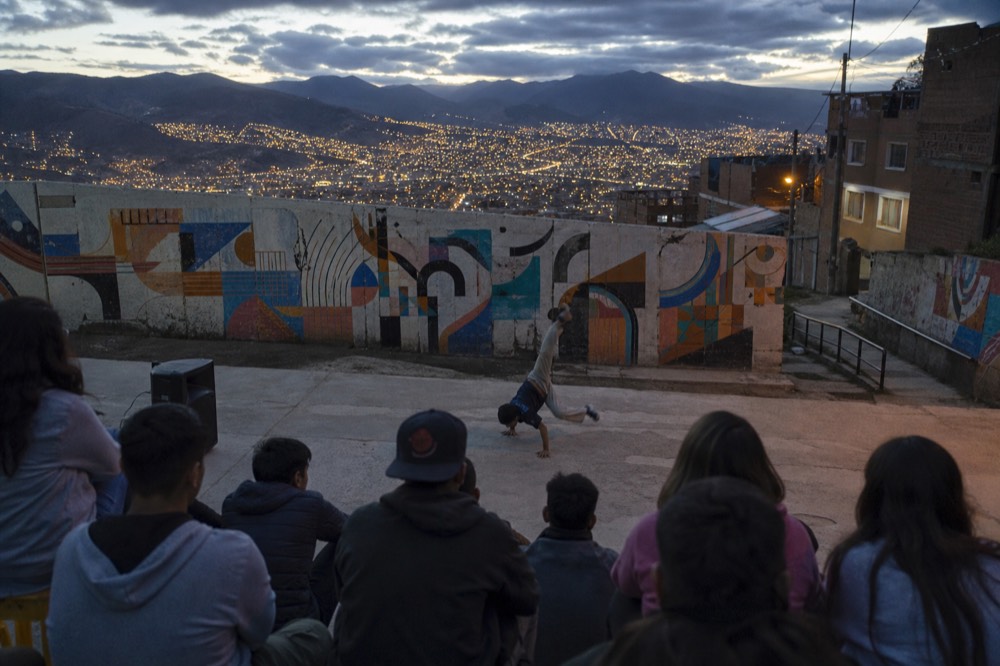 A breakdancing performance on a concrete landing with a ring of onlookers seen from behind, speaker on the ground, abstract mural wall as backdrop