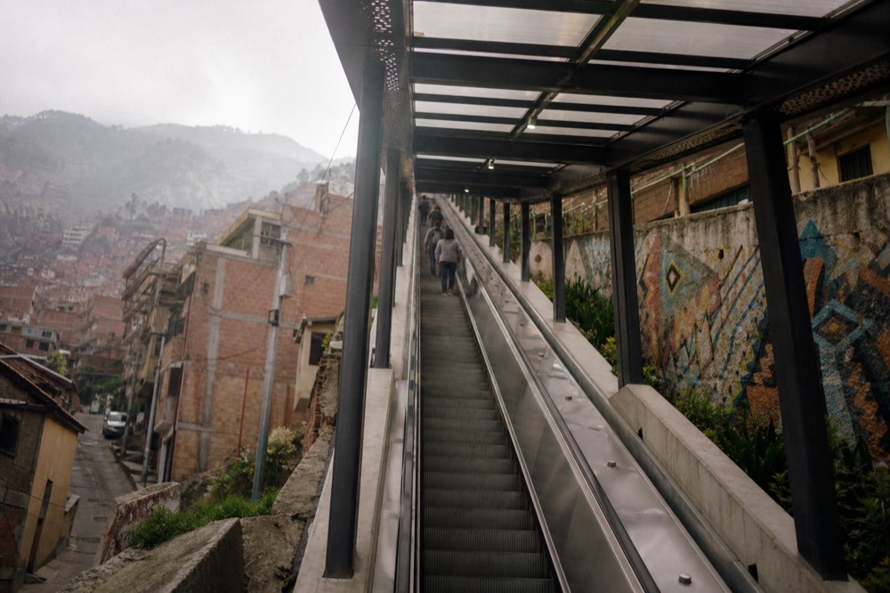 Covered escalator flight photographed from below, ribbed metal steps rising steeply under a bright canopy roof with brick houses on both sides