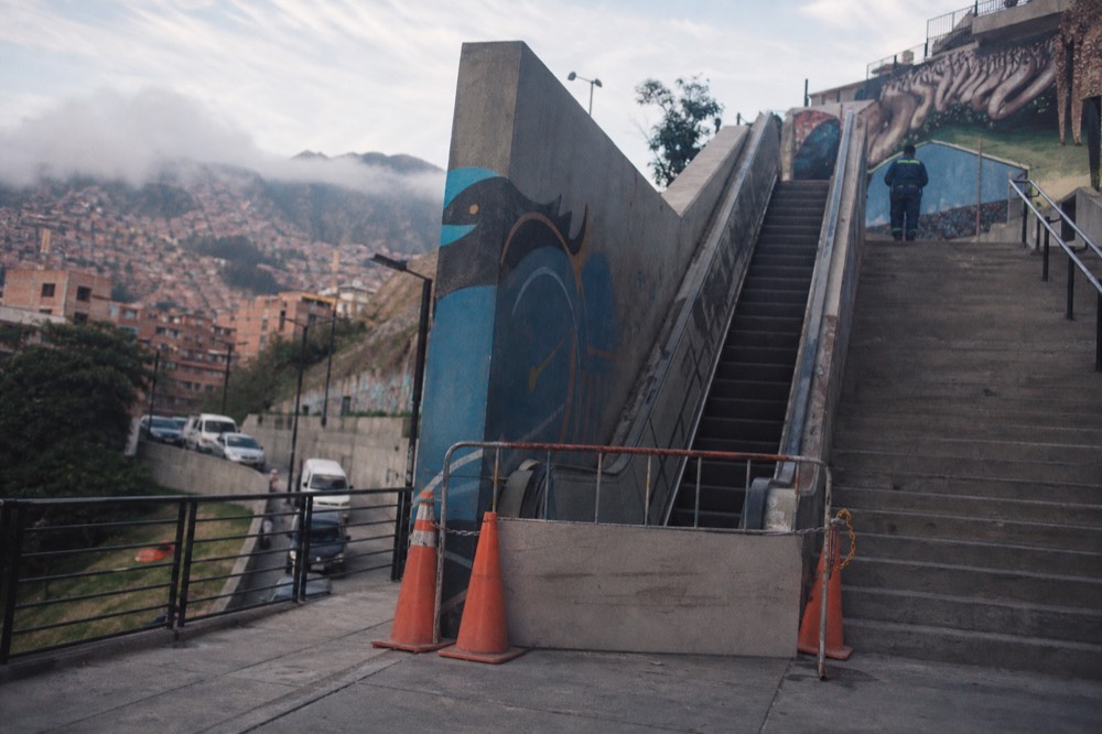 Section of escalator closed behind plain barriers and traffic cones with a distant worker in overalls, adjacent concrete stairs still open