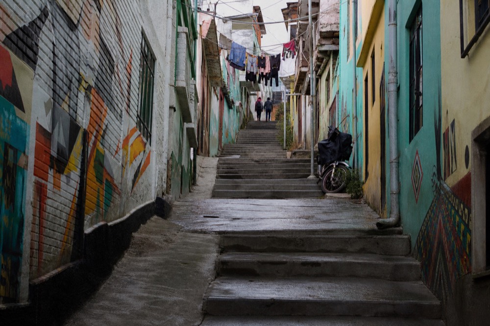 Narrow concrete stair alley climbing steeply between painted brick houses with laundry lines overhead and two distant figures walking up