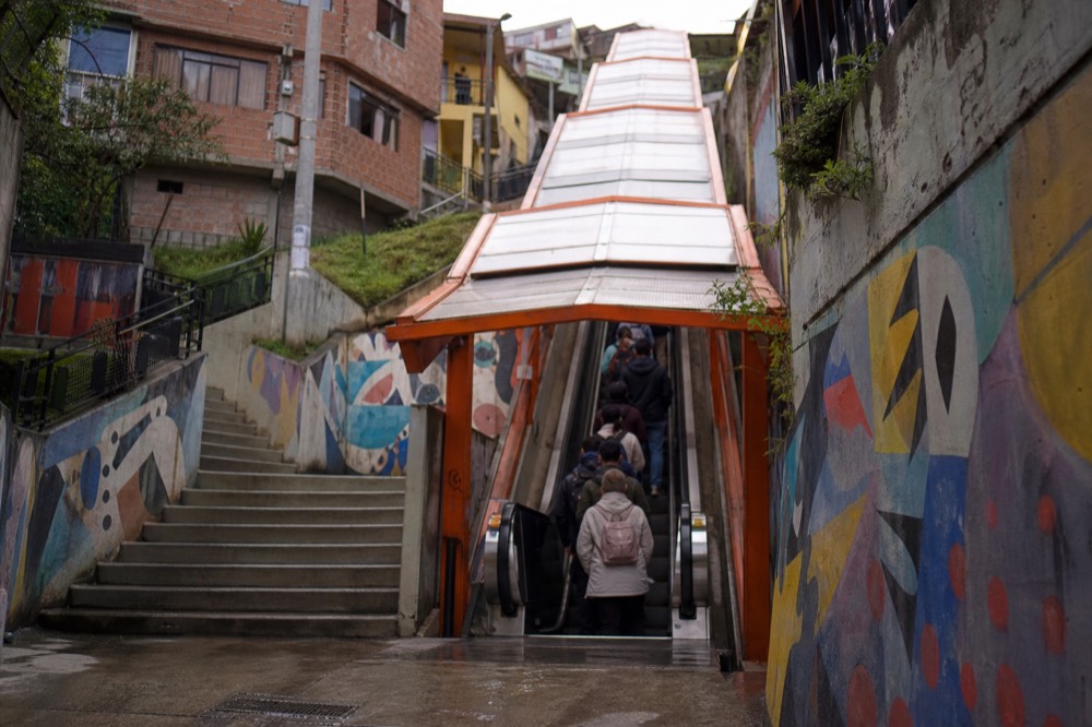 Outdoor public escalator with an orange canopy climbing a steep hillside between brick houses, riders visible from behind on the moving steps