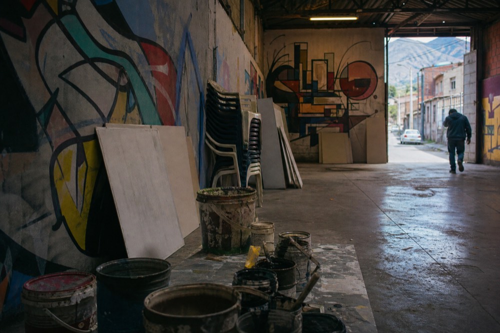 Small community arts room with stacked plastic chairs, open paint cans and blank primed boards, afternoon light coming through an open doorway
