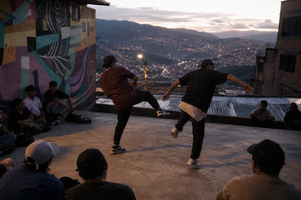 Two street dancers mid-move on a concrete landing ringed by seated onlookers, painted wall behind them, late afternoon shadows