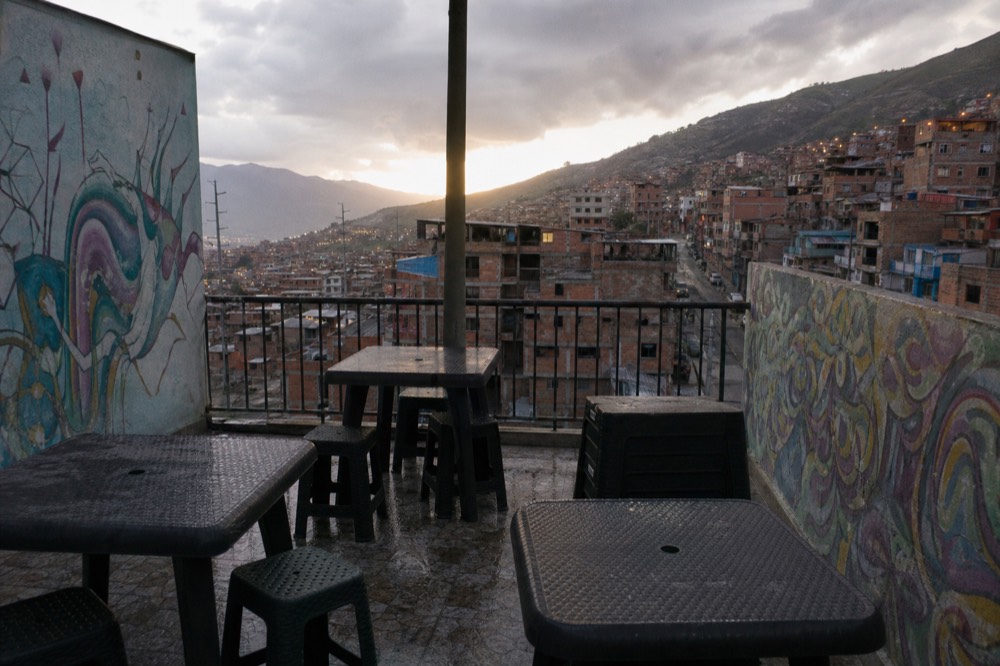 Rooftop terrace with plastic tables and a metal railing overlooking a dense valley of brick houses at golden hour