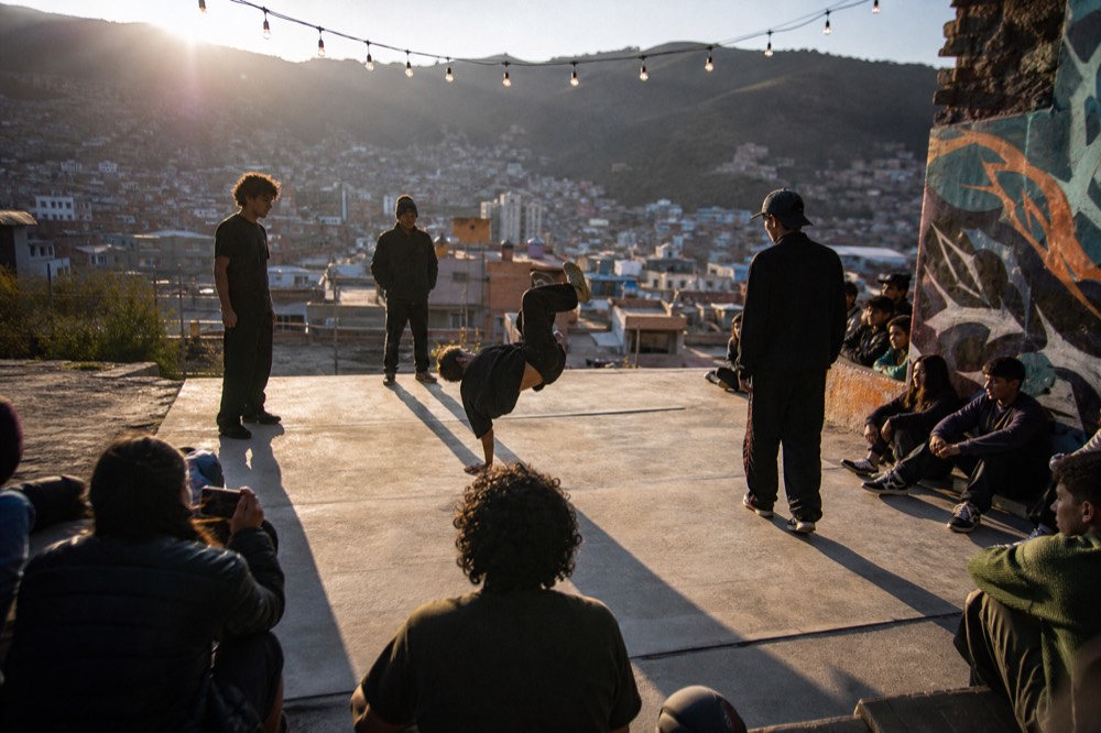 Group of breakdancers performing on a concrete platform ringed by seated onlookers, low sun casting long shadows across the ground