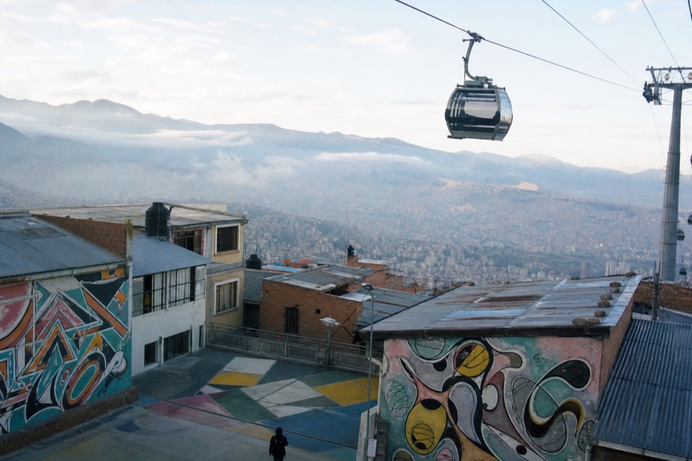 Cable-car cabin gliding above corrugated rooftops and painted courtyards with valley haze stretching into the distance