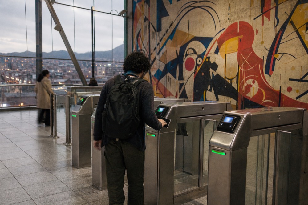 Metro station ticket gates with a passenger tapping a card at the barrier, tiled floor and daylight through the glass wall behind