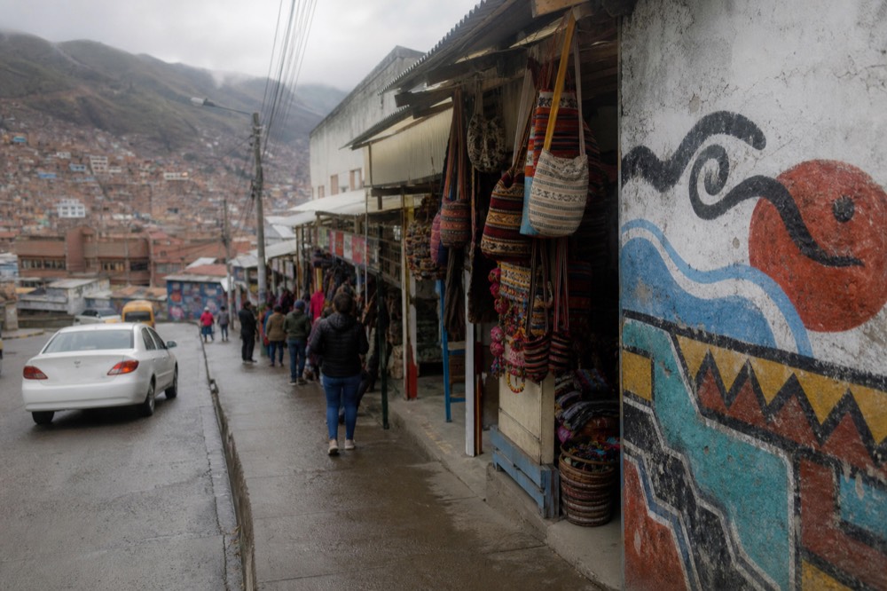 Row of open-fronted craft stalls hung with woven bags and bright textiles along a narrow terrace walkway, shoppers browsing at a distance