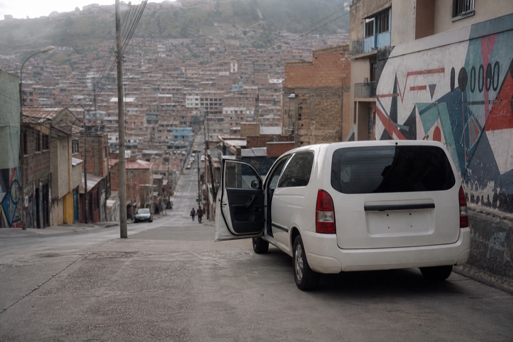 Unmarked white minivan waiting on a steep residential street with the driver's door open, hillside houses rising behind