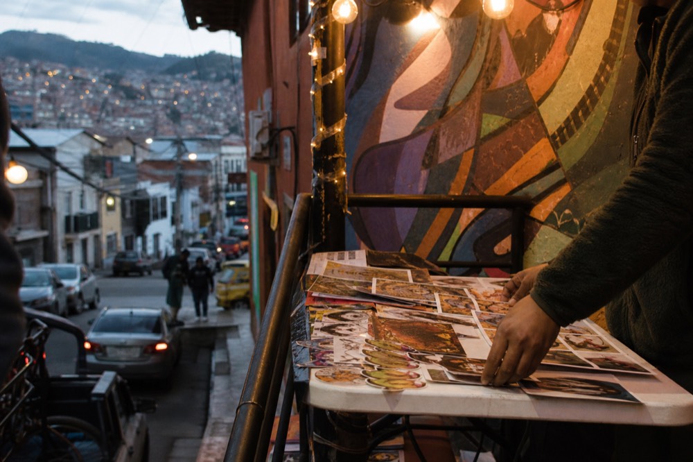 Small prints and stickers laid out on a folding table at a street stall, a seller's hands arranging them, painted wall behind