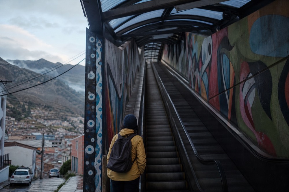 Solo traveller with a small backpack standing at the foot of a covered outdoor escalator, looking up the steep painted corridor, no crowd around