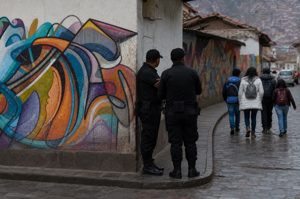Two police officers standing at a corridor junction beside a mural-covered wall while visitors pass by in daylight, calm ordinary scene