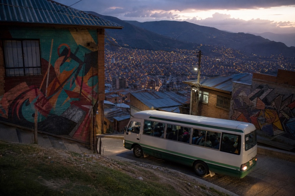 Small local minibus climbing a narrow steep street lined with brick houses and shopfronts, passengers visible through the windows, hillside rising behind