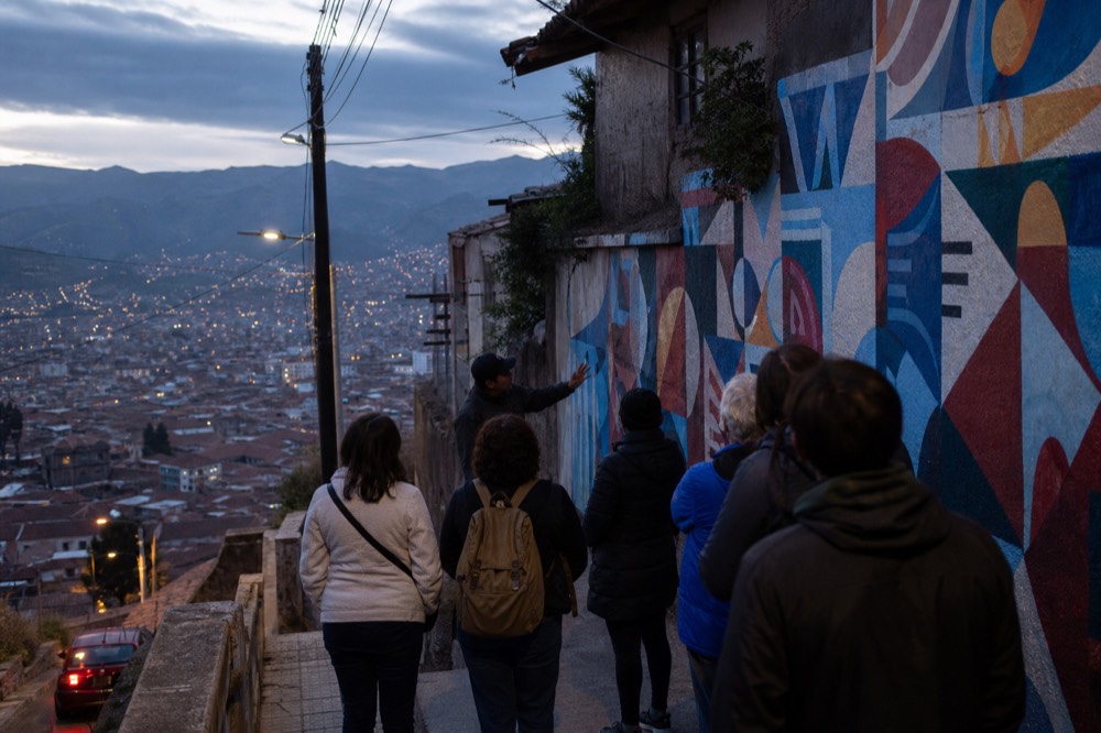 A guide gesturing toward a large abstract mural while a group of six visitors listens, seen from behind on a narrow hillside walkway in soft light