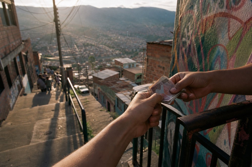 Close view of hands passing folded Colombian banknotes at a hillside terrace railing, blurred valley of red-brick houses falling away behind