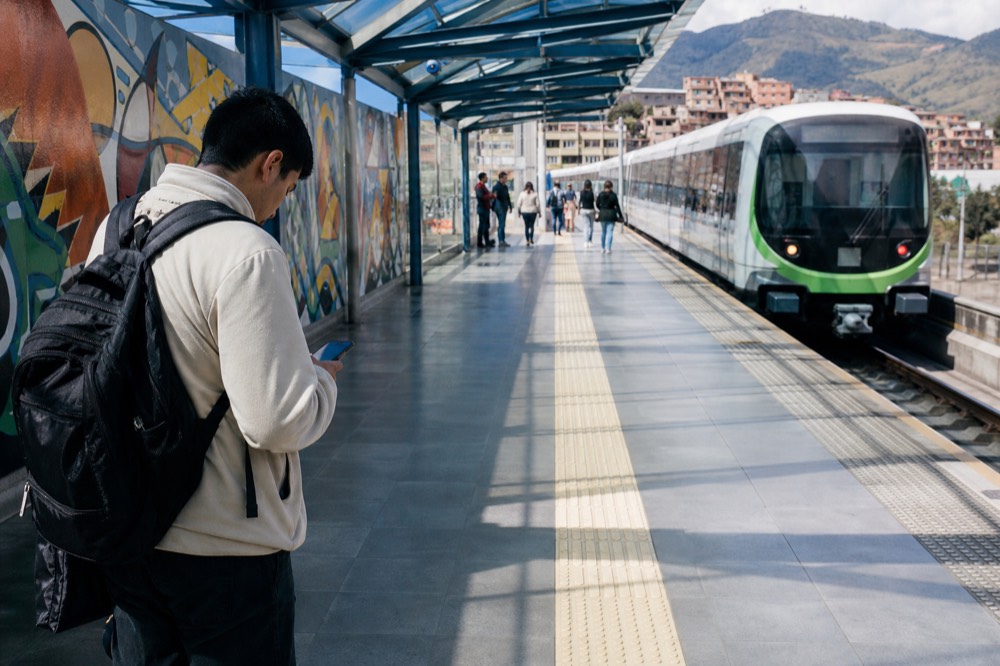 Traveller checking a phone screen on a bright metro platform with a train arriving, tiled floor and glass canopy overhead, other passengers blurred