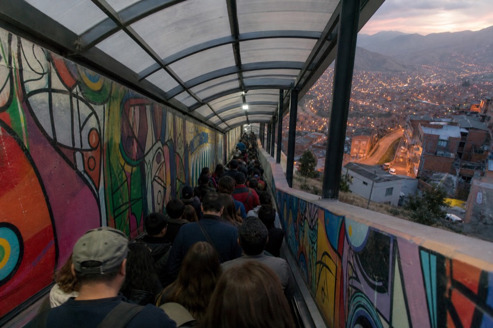 Dense crowd of visitors packed onto a covered outdoor escalator on a steep hillside, painted walls either side, bright midday light