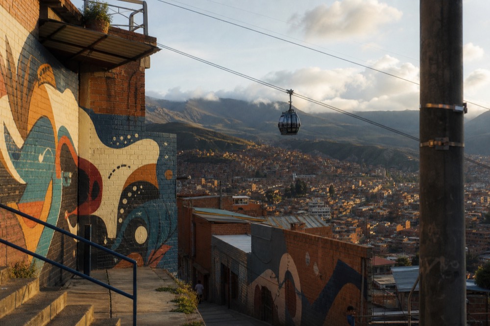 A glass cable car cabin rising above a hillside of dense red-brick homes, mountains and low cloud filling the valley behind it