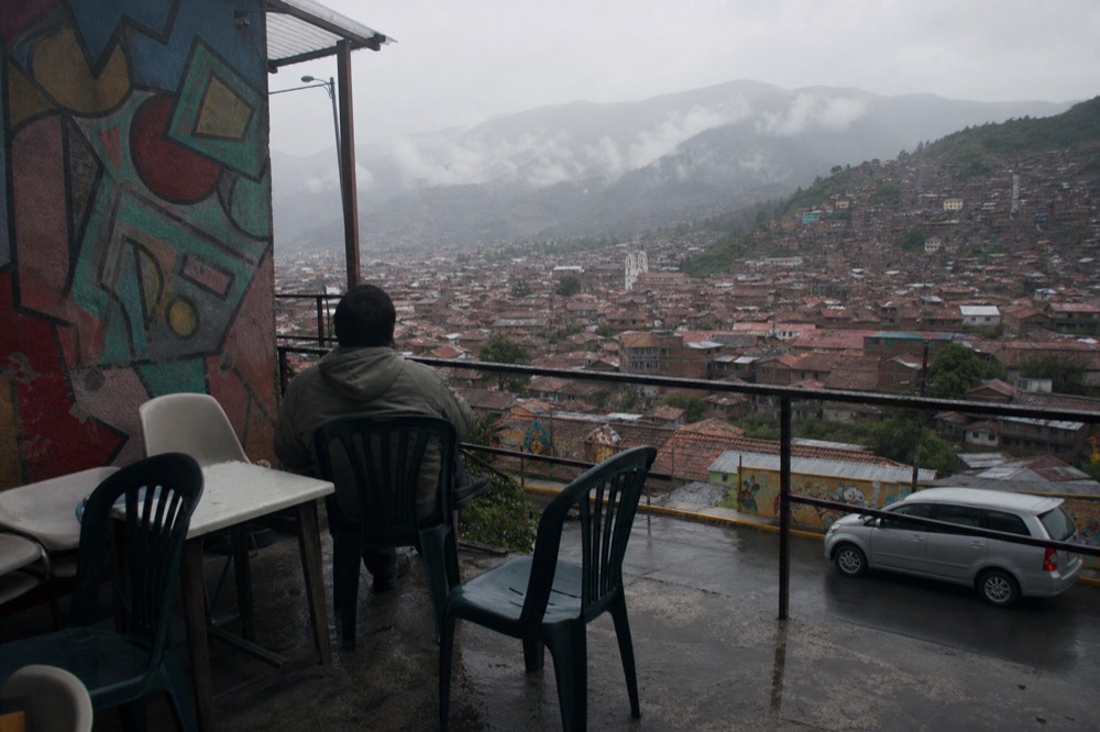 A hillside terrace cafe with plastic chairs and a low railing, one visitor seated looking out over a wide valley of tightly packed rooftops