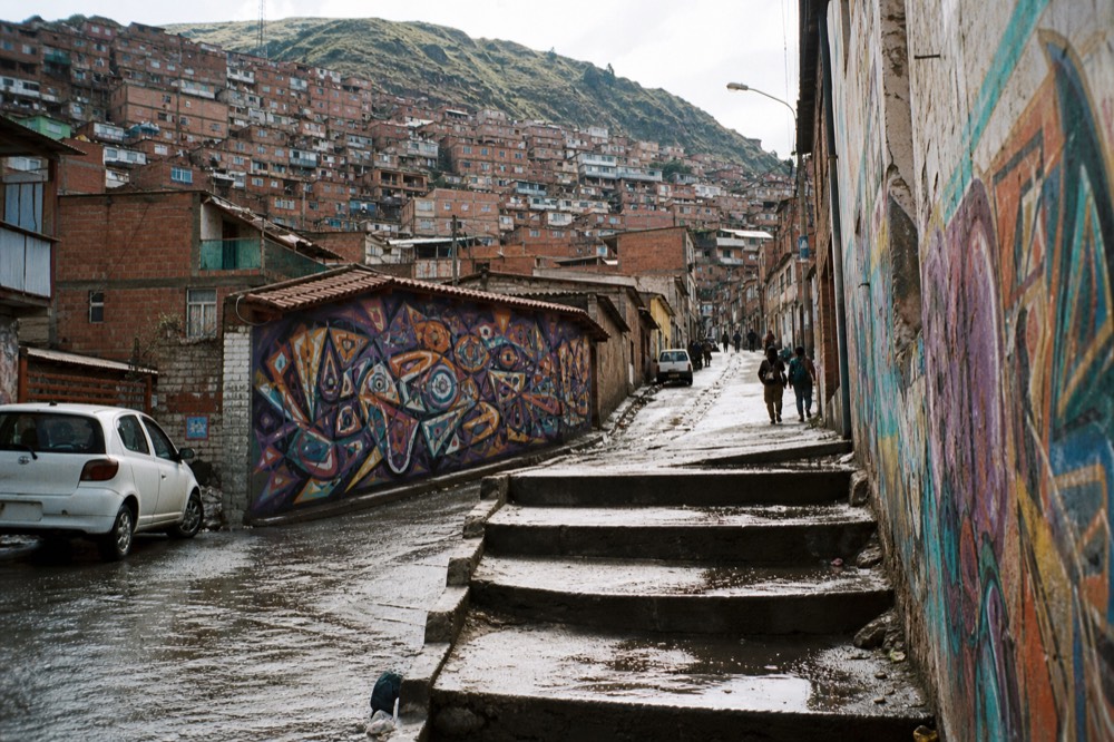 Steep hillside covered in stacked red-brick houses with corrugated roofs rising above a busy street, distant figures walking up a narrow lane