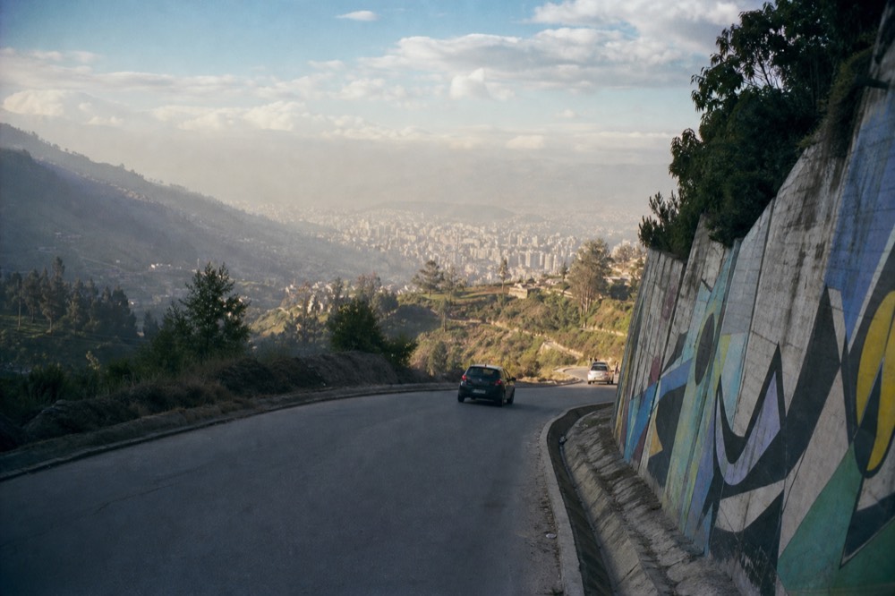 Winding mountain highway descending through green ridges into a hazy valley of city buildings under a soft afternoon sky