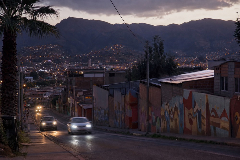 City avenue at dusk with car headlights streaking past low buildings and palm trees, mountain silhouette and lit hillside houses behind