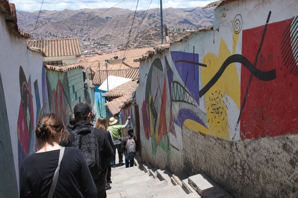 Small group of visitors seen from behind following a guide up a narrow stairway alley lined with colourful abstract painted walls
