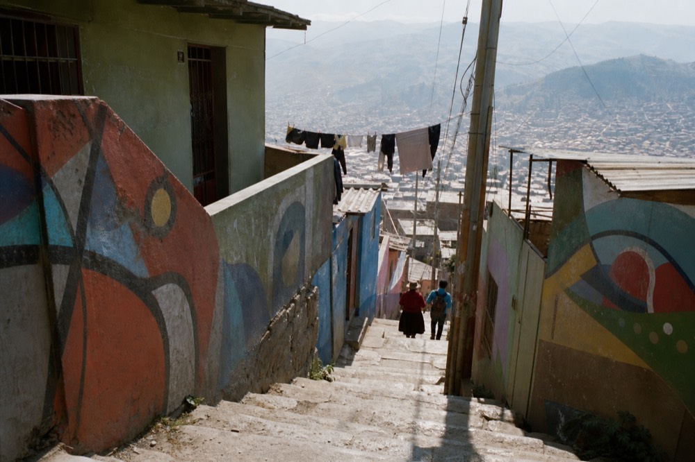 A narrow hillside stairway alley between painted concrete houses, washing lines overhead, two distant figures climbing away from the camera in bright midday light